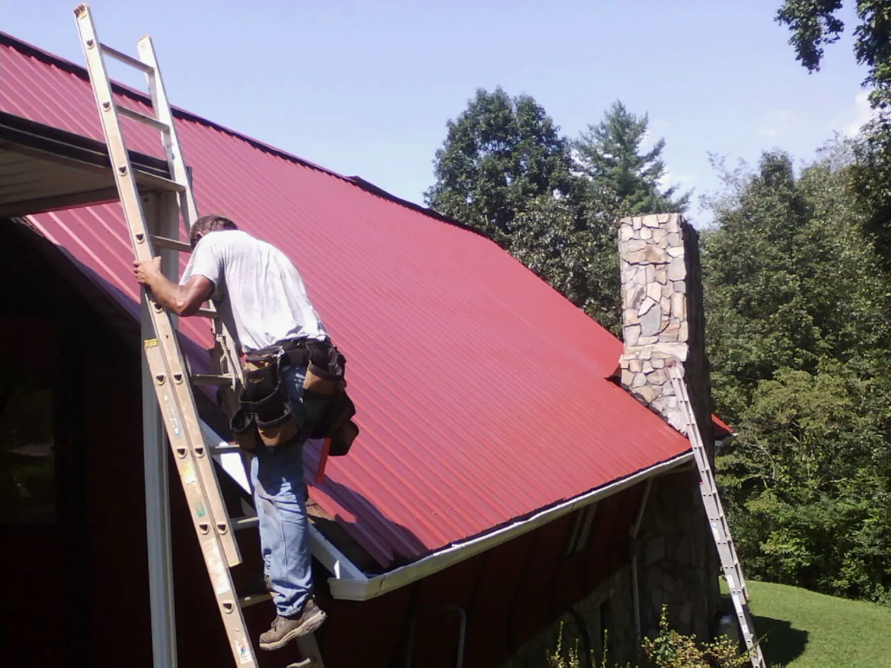 Roofer on a ladder beside a red metal roof and stone chimney — Soffit & Fascia Repair by Roofing Brothers in Farmdale